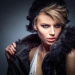 A striking fashion portrait of a young woman with blonde hair, wearing a fur-lined vest and hat, looking intensely at the camera against a moody grey background.