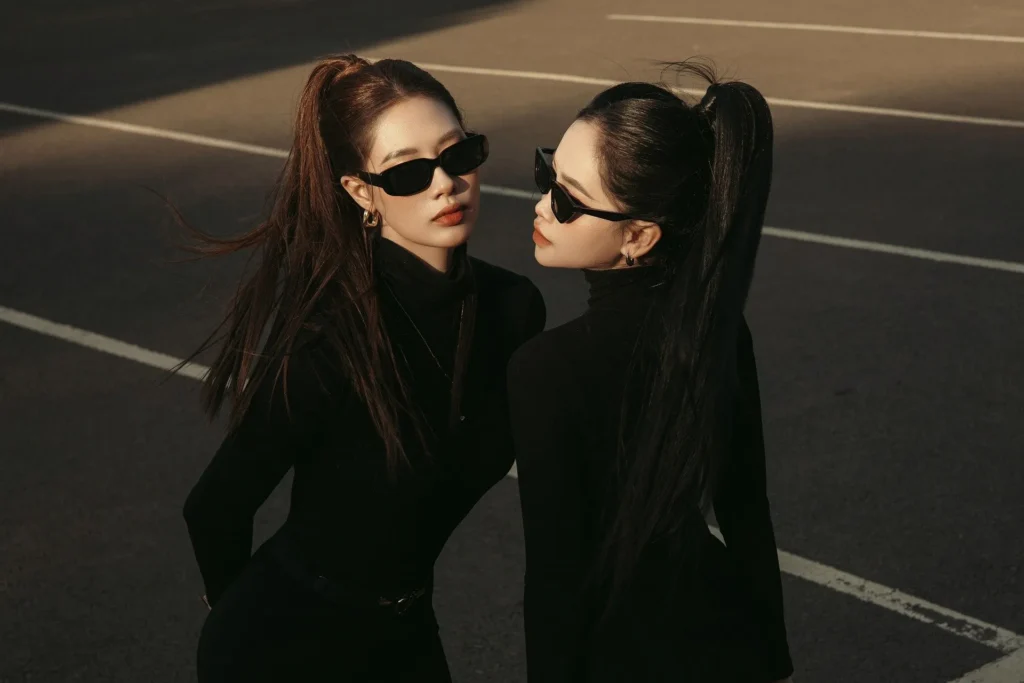Two fashionable women with long high ponytails and black sunglasses pose in matching black turtlenecks against a minimalist asphalt background with harsh, cinematic lighting.