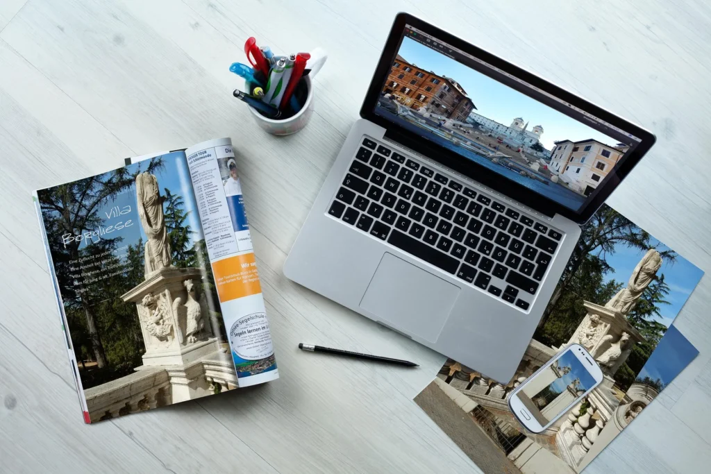 A top-down view of a modern workspace featuring a laptop displaying a scenic photo of Rome, a travel magazine opened to a "Villa Borghese" article, a smartphone, and printed photos, symbolizing the blend of digital and traditional vacation planning.