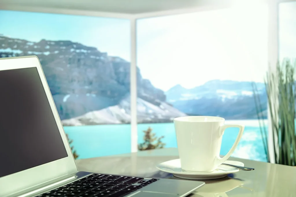 A laptop on a desk next to a white coffee cup, displaying a blurred view of a turquoise mountain lake through a window, illustrating how AI travel planning tools help users book scenic vacations from home.