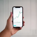 A hand holding a smartphone displaying a vertical cryptocurrency candlestick chart with green and red bars and technical indicators for crypto day trading.