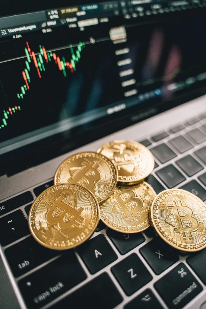 Five gold Bitcoin coins resting on a laptop keyboard in front of a digital screen displaying a green and red candlestick price chart.