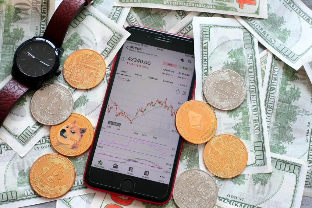 Day Trading Cryptocurrency Concept on a smartphone displaying a Bitcoin candlestick price chart surrounded by physical cryptocurrency coins (Bitcoin, Ethereum, Dogecoin), USD banknotes, and a wristwatch.