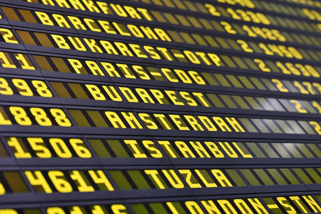 A close-up, angled view of a digital airport departure board displaying flight destinations like Barcelona, Paris, Budapest, and Amsterdam in glowing yellow text.